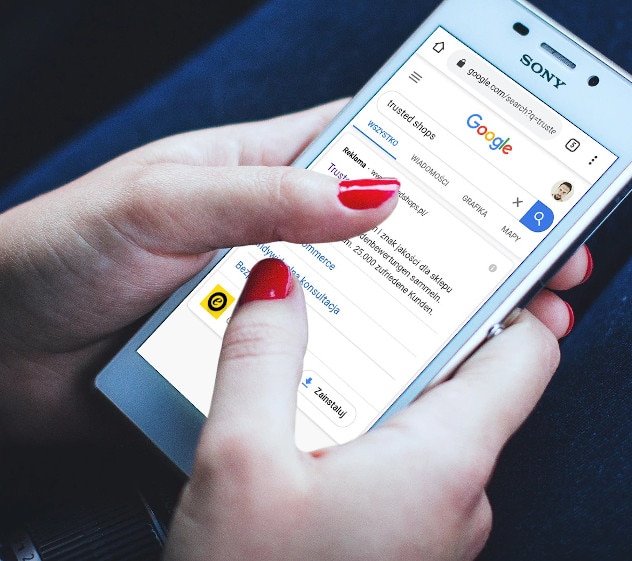 Na obrazku widać dłoń operującą smartfonem. Na smartfonie wyświetla się wynik wyszukiwania z reklamą Trusted Shops.