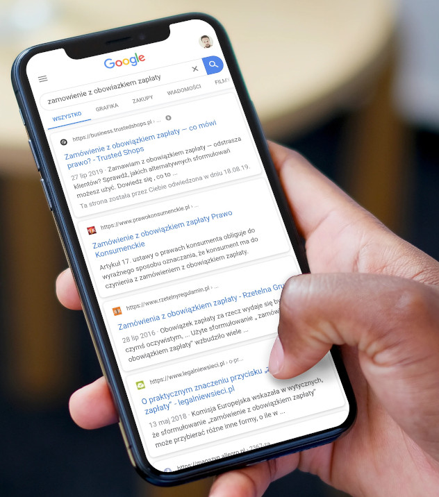 Na obrazku widać dłoń operującą smartfonem. Na smartfonie wyświetla się przykładowy wynik wyszukiwania. Obok wyników wyszukiwania można dostrzec różne favicony.